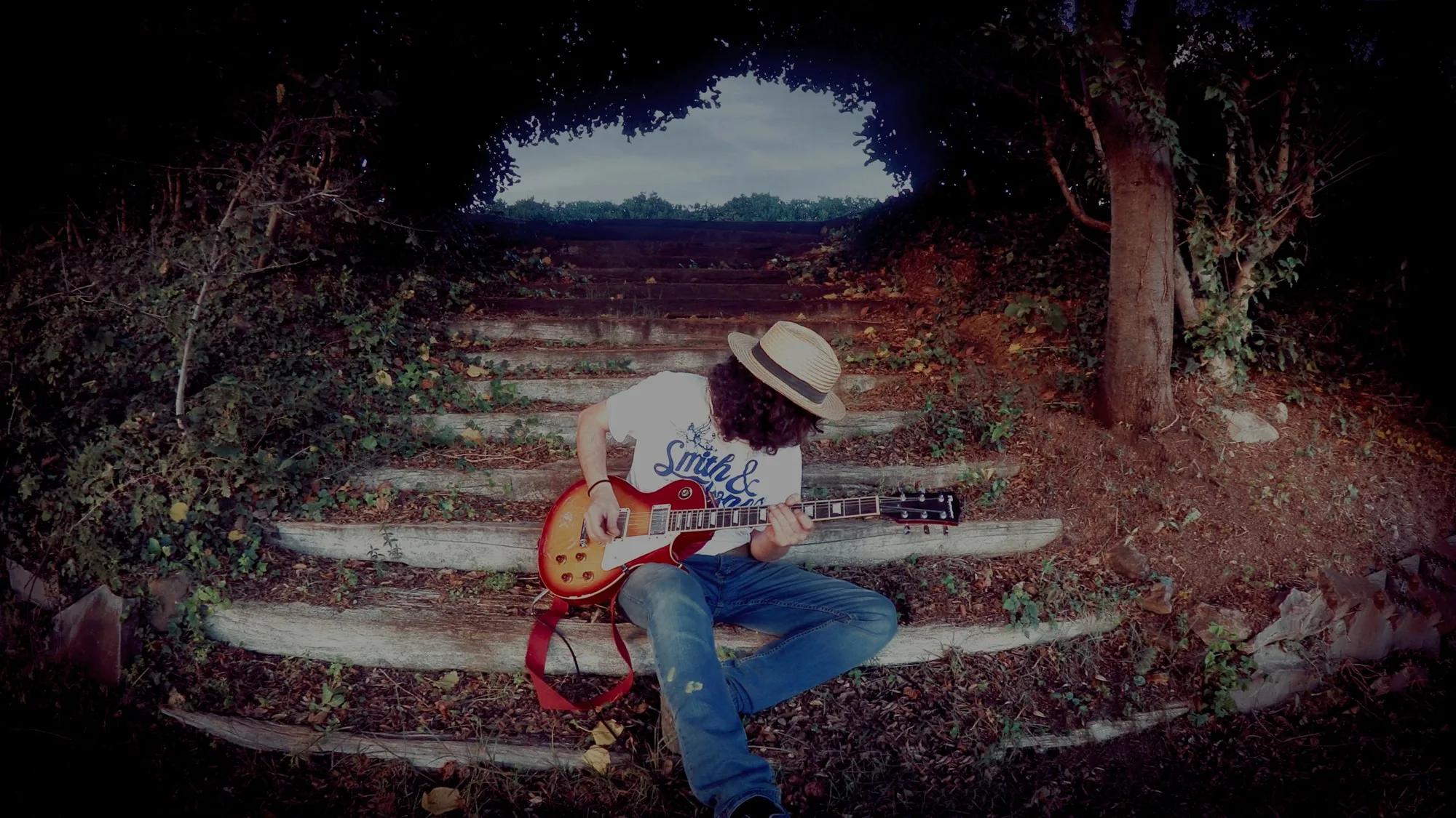 A man with curly dark hair and a straw hat sits on worn stone steps inside a tunnel of trees, playing a sunburst electric guitar, with a patch of sky showing through the opening above.